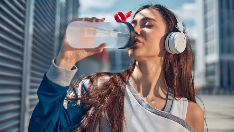 Young woman with headphones drinking water after workout. Outdoor city background, sunny day.